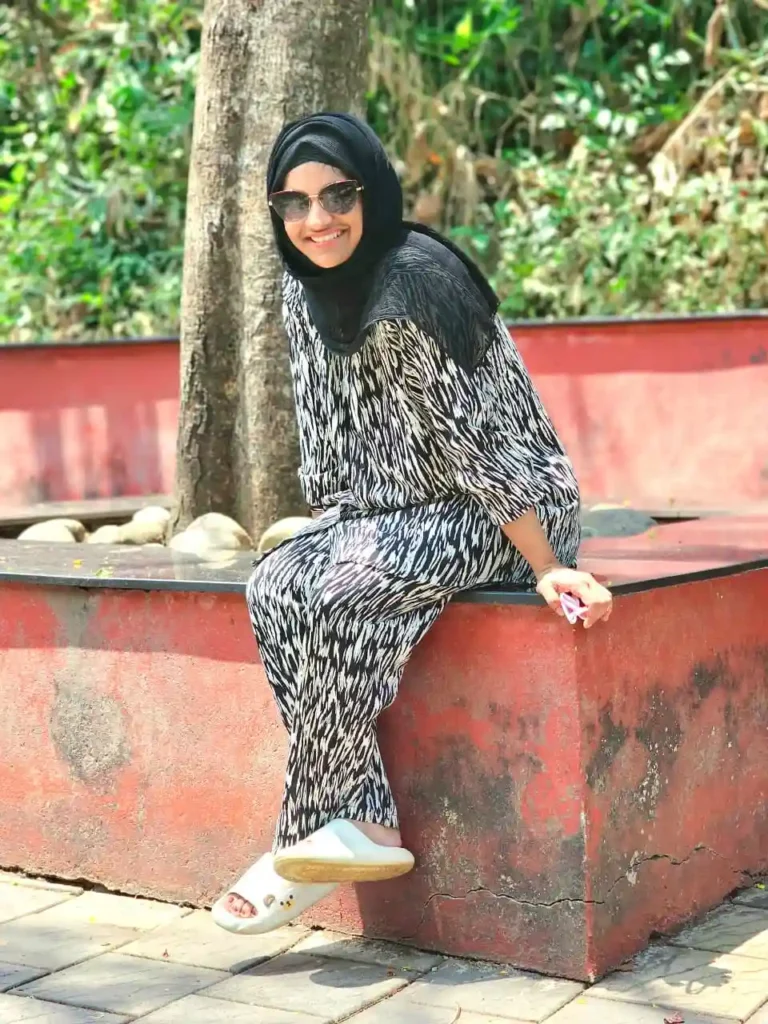 One of the Best Digital Marketer in Thrissur Faseela Nahas wearing a black headscarf and sunglasses, sitting outdoors near a tree in a relaxed setting